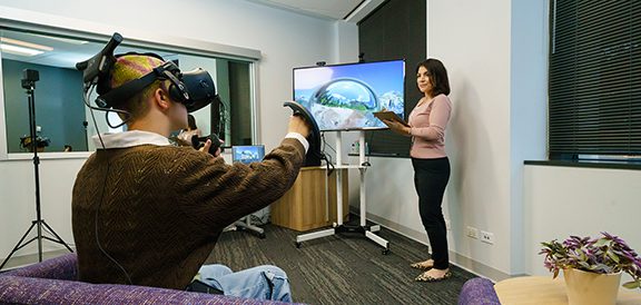 A person wearing a virtual reality headset and using controllers participates in a UX research session, guided by a researcher standing next to a large screen displaying a virtual environment.